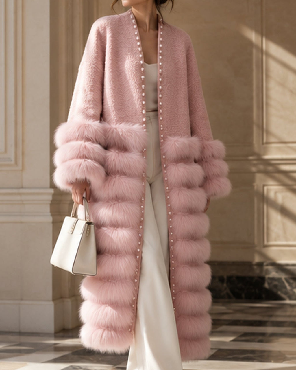 A woman wearing a long, fluffy pink faux-fur coat with oversized matching cuffs, styled with white pants, a white top, and a white handbag in an elegant indoor setting.