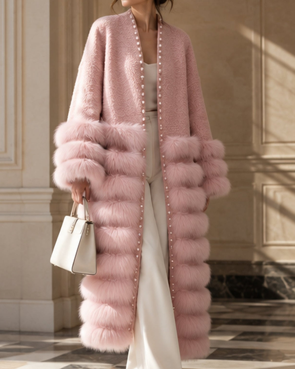 A woman wearing a long, fluffy pink faux-fur coat with oversized matching cuffs, styled with white pants, a white top, and a white handbag in an elegant indoor setting.