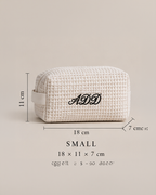 A small textured cream-colored cosmetic pouch with black monogrammed initials 'ADD' on the front, shown with dimension lines indicating measurements of 18 cm by 11 cm by 7 cm, on a solid beige background.