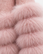 A close-up of a soft pink faux fur garment with layered, fluffy pom-pom style texture and a plush lining visible at the top, shown against a plain light background.