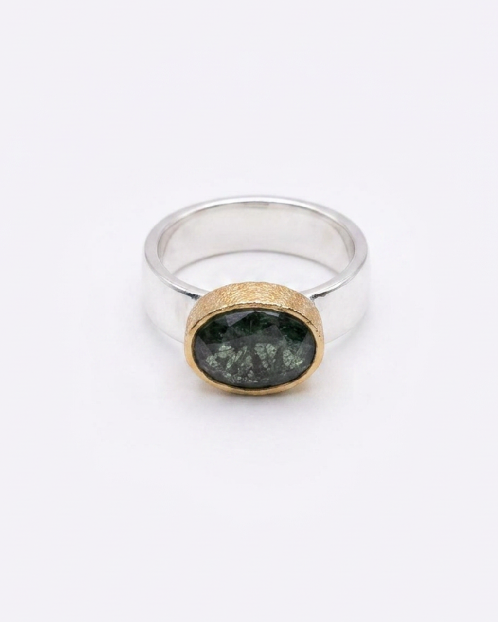 A silver ring with a textured gold bezel holding a dark green, speckled oval stone, shown on a plain white background.