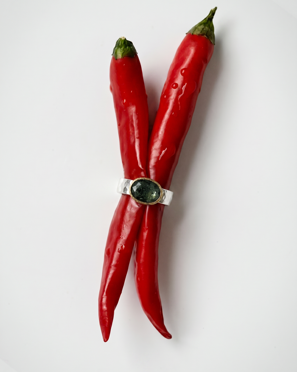 A silver ring with a round dark green stone, wrapped around two red chili peppers on a plain white background.