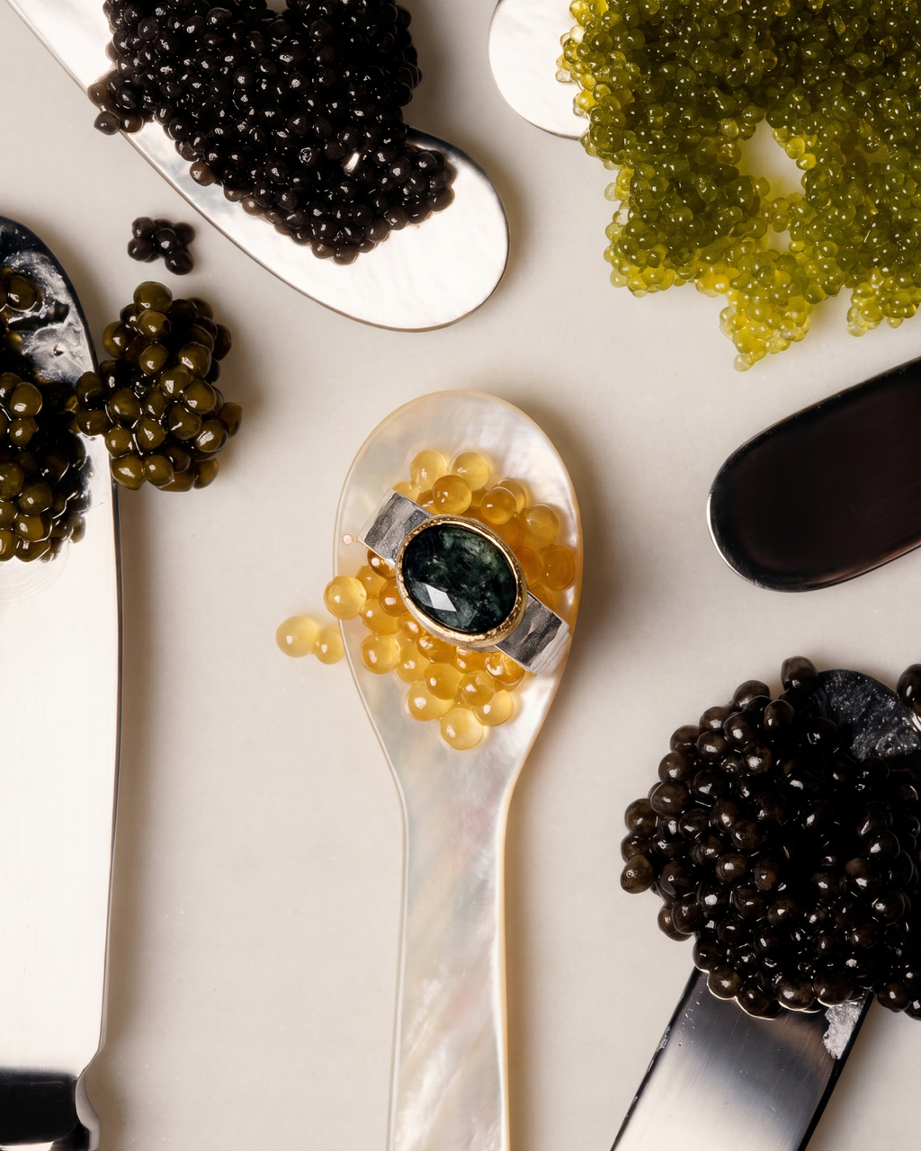 A close-up flat lay of caviar on spoons, with a central spoon holding amber-colored caviar and a dark green oval gemstone ring set in silver. Other spoons contain black, olive-green, and yellow caviar on a light beige surface.
