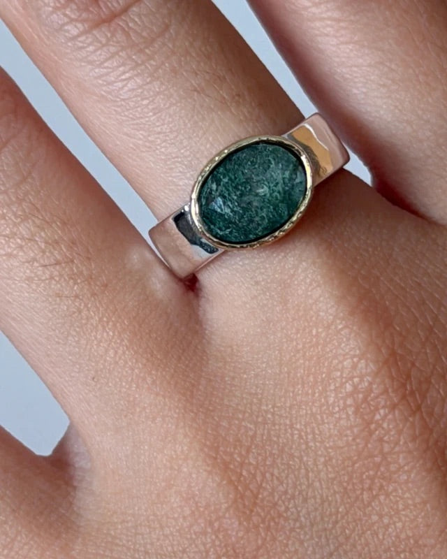 A close-up of a hand wearing a silver ring with an oval green stone set in a gold-toned bezel.