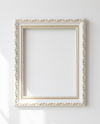 An ornate white picture frame with gold decorative detailing, shown hanging on a plain white wall with soft sunlight and shadows.