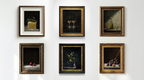 Six framed still life paintings arranged in two rows on a white wall. The paintings feature dark backgrounds with various objects including a gift box, wine glasses, a bottle, fruit, and flowers, each in different ornate frames.