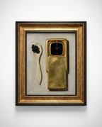 A framed artwork on a white wall featuring a sculptural still life: a gold-toned rectangular object with a dark face, a cream spoon with dark berries, and a textured gray background, all enclosed in a gold and black frame.