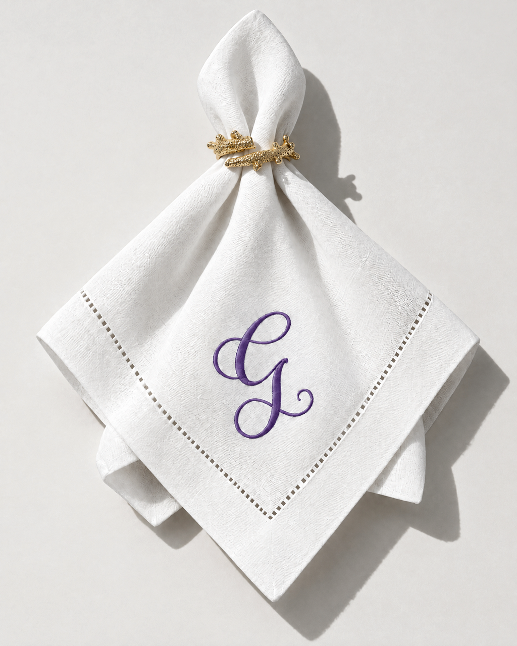 The Soirée Napkin with a purple embroidered initial, gathered in a gold napkin ring