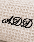 Close-up of a cream waffle-weave fabric item, likely a towel or blanket, with black embroidered cursive initials 'ADD' on the surface.