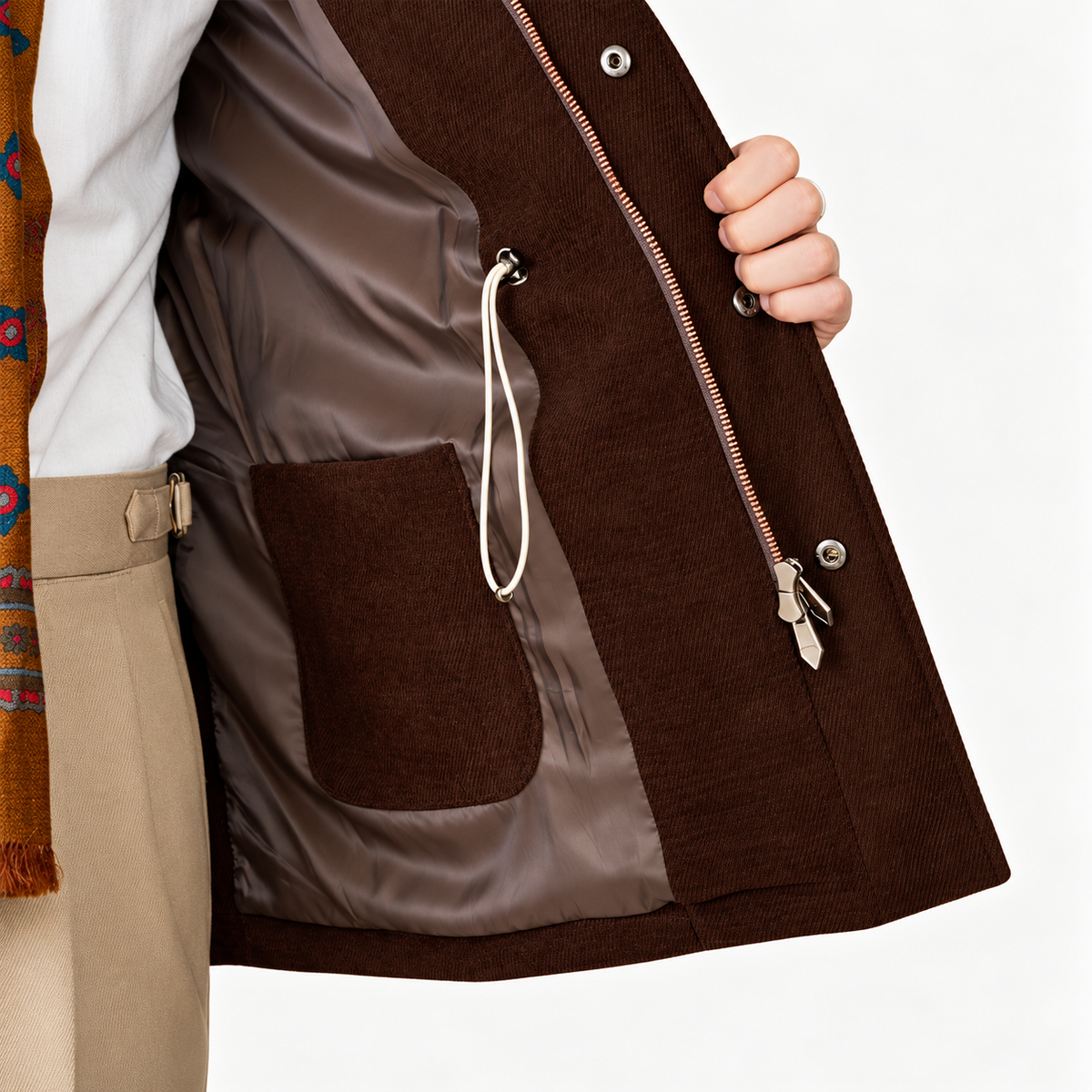 Close-up of a person wearing a dark brown corduroy jacket with a satin brown lining, a front zipper, a drawstring pocket, and a large patch pocket, paired with beige trousers and a patterned scarf.