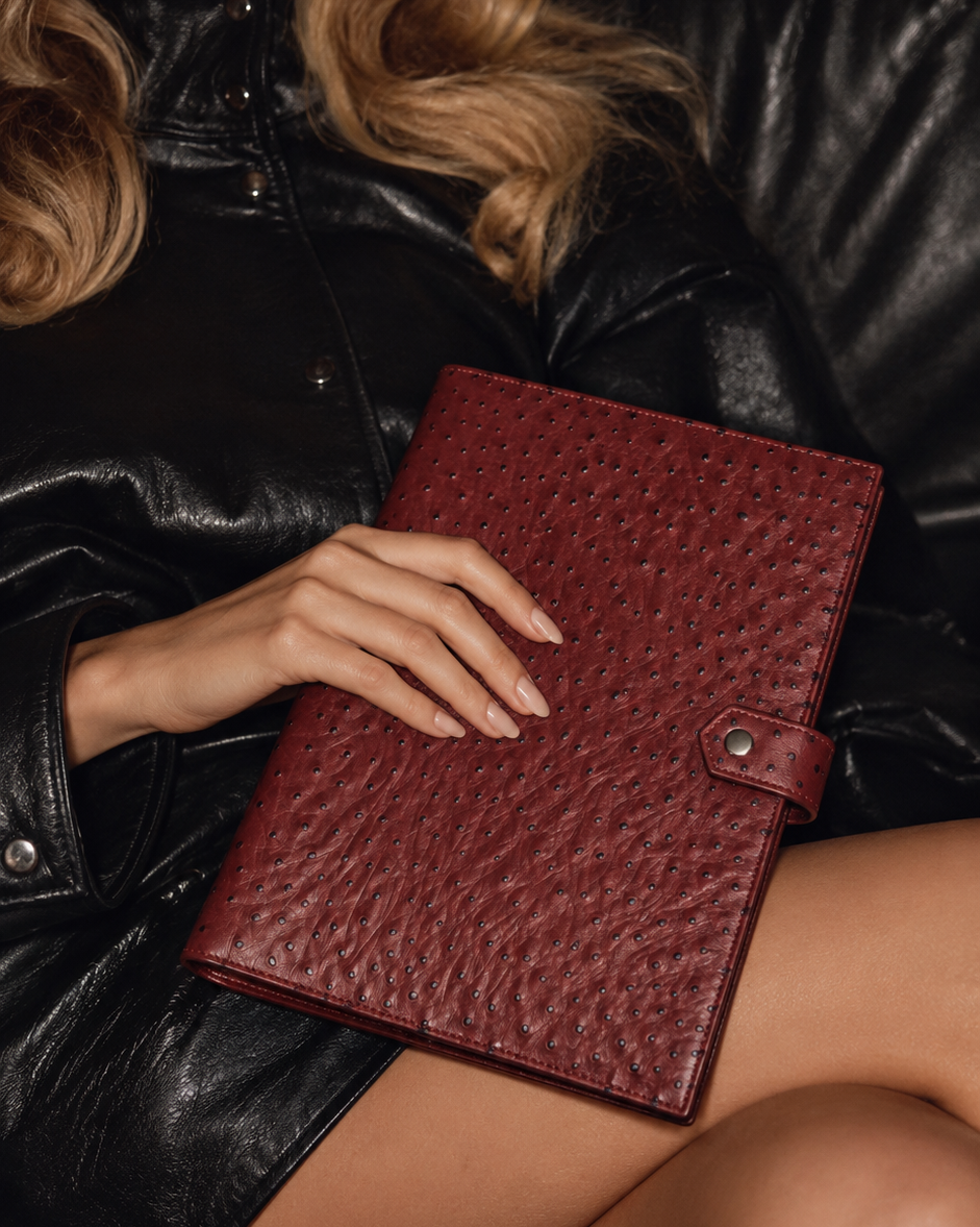 A woman in a black leather outfit holds a burgundy textured leather planner or folder with a strap closure, seated on a black leather couch.