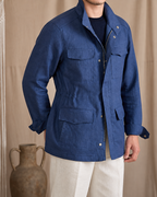 A man wearing a blue linen utility jacket over a black shirt and cream pants, standing in front of a beige curtain with a macrame wall hanging and a rustic vase in the background.