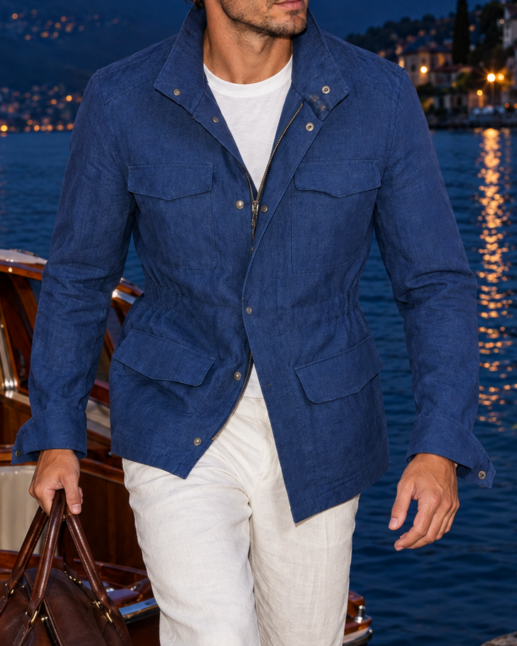 A man wearing a blue textured jacket and white trousers walks along a stone pier next to a lake at dusk, holding a brown leather duffel bag. A wooden boat is docked on the left, and a lit town and mountains are visible in the background.