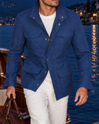 A man wearing a blue textured jacket and white trousers walks along a stone pier next to a lake at dusk, holding a brown leather duffel bag. A wooden boat is docked on the left, and a lit town and mountains are visible in the background.