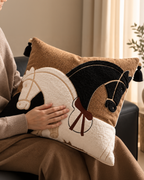 A person holding a square decorative throw pillow with an embroidered horse design in brown, cream, and black, set in a cozy indoor environment.