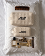 Two cream waffle-knit cosmetic pouches with black embroidered monogram 'ADD' are arranged on a white pillow with other luxury items including a YSL lipstick, YSL perfume bottle, and a comb.
