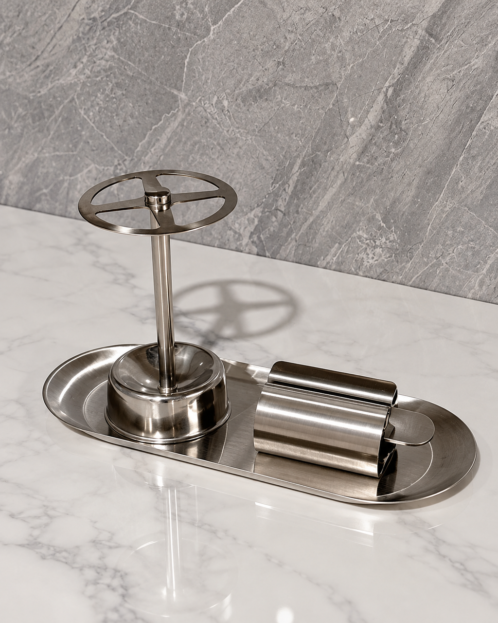 A brushed stainless steel bathroom accessory set on a white marble surface, featuring a wall-mounted faucet handle with a wheel design, a matching cylindrical holder, and a small oval tray.