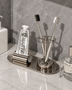 A stainless steel bathroom organizer set on a white marble countertop against a gray marble backsplash. The set includes a toothbrush holder with two toothbrushes, a toothpaste tube holder with a tube of Marvis toothpaste, and a small tray.
