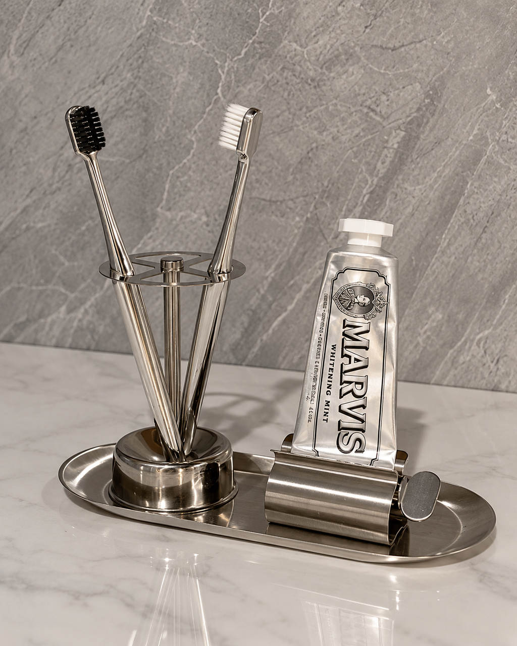A stainless steel bathroom organizer set on a marble countertop, featuring a toothbrush holder with two toothbrushes, a squeeze tube of MARVIS toothpaste, and a toothpick holder on an oval tray.
