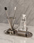 A stainless steel bathroom organizer set on a marble countertop, featuring a toothbrush holder with two toothbrushes, a squeeze tube of MARVIS toothpaste, and a toothpick holder on an oval tray.