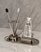 A stainless steel bathroom organizer set on a marble countertop, featuring a toothbrush holder with two toothbrushes, a squeeze tube of MARVIS toothpaste, and a toothpick holder on an oval tray.