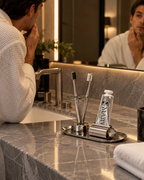 A man in a white bathrobe looks into a bathroom mirror while grooming. On the gray marble countertop in front of him is a silver toothbrush holder with two toothbrushes, a tube of toothpaste labeled DAILY, and a small metal cup.