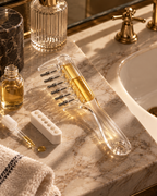 A clear plastic and glass hair styling tool with multiple metal bristles, shown on a marble bathroom counter beside a sink, gold faucet, small dropper bottle, and a white applicator tray.