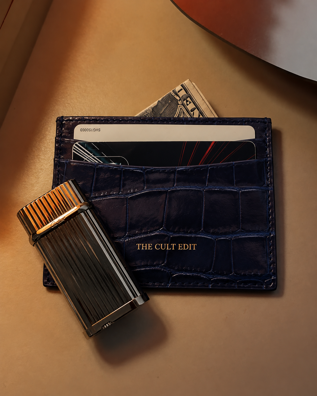 A dark navy blue crocodile-embossed leather card holder wallet with gold lettering reading 'THE CULT EDIT', shown with cards and cash partially inserted, alongside a metallic black and gold lighter on a warm beige surface.