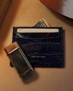 A dark navy blue crocodile-embossed leather card holder wallet with gold lettering reading 'THE CULT EDIT', shown with cards and cash partially inserted, alongside a metallic black and gold lighter on a warm beige surface.