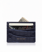 A navy blue crocodile-embossed cardholder wallet with gold 'THE CULT EDIT' branding, shown with credit cards and a dollar bill partially inserted, on a white background.