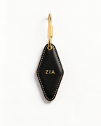 A black leather keychain with a diamond-shaped body, white contrast stitching, and gold-tone hardware. The front of the leather piece is personalized with the name 'ZIA' in gold lettering.