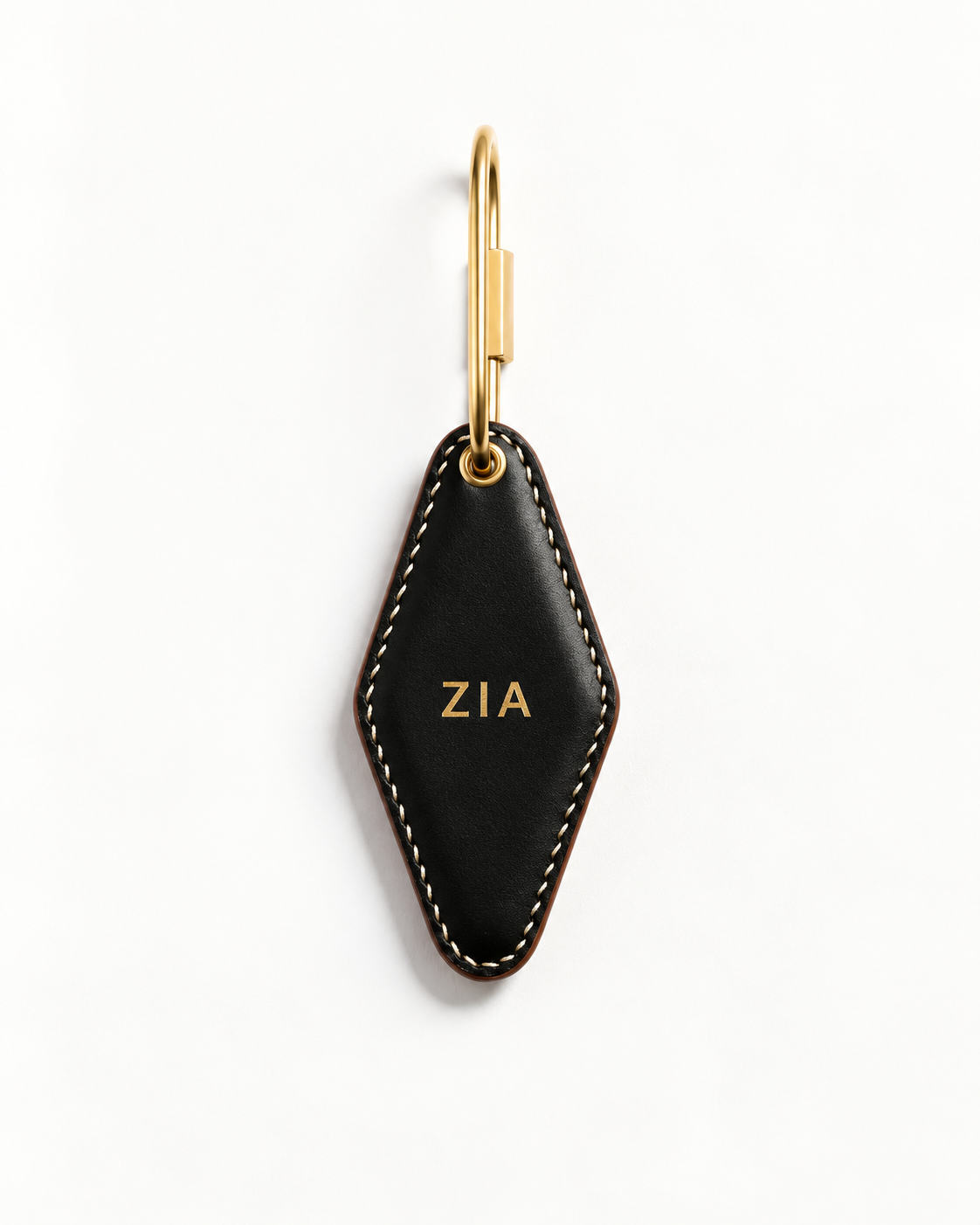 A black leather keychain with a diamond-shaped body, white contrast stitching, and gold-tone hardware. The front of the leather piece is personalized with the name 'ZIA' in gold lettering.