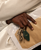 A close-up of a person's hand holding a set of keys with a dark green leather keychain featuring the gold initials 'ZIA', resting on a beige textured surface next to a clear plastic bag.