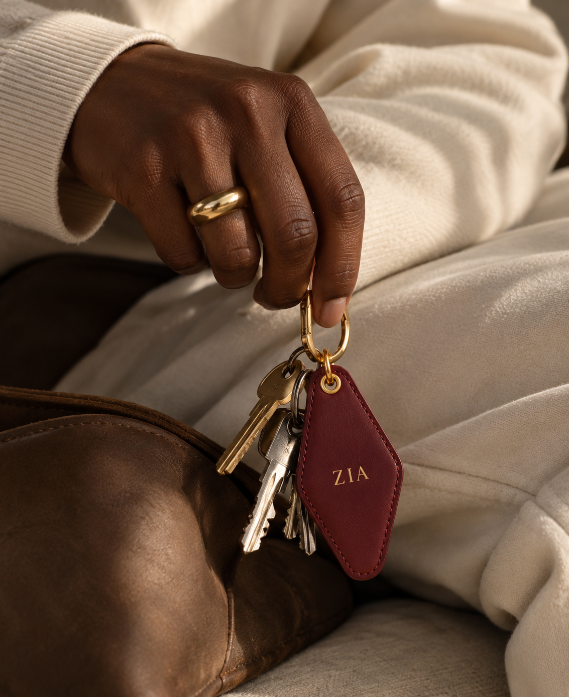 A close-up of a person's hand holding a set of keys with a personalized burgundy leather keychain featuring gold hardware and the initials 'ZIA'.