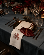An elegantly styled dinner table setting with a slate blue tablecloth, gold-toned cutlery, clear wine glasses, and a white napkin featuring a red embroidered monogram letter 'M' with floral detailing.