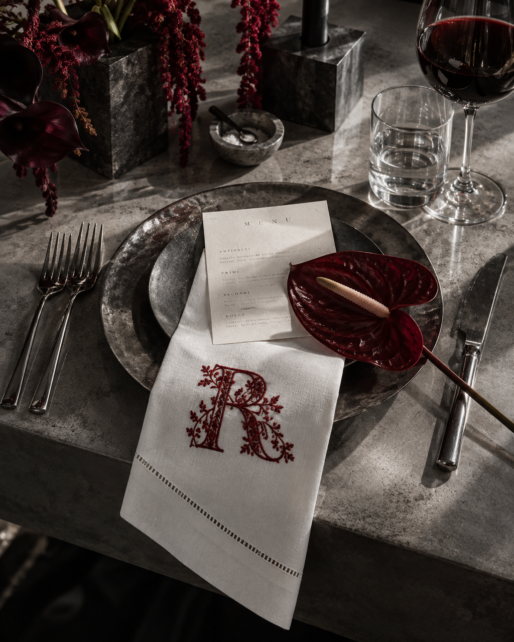 A styled dining table setting featuring a white linen napkin with an embroidered red monogram letter 'R' and floral border, layered over a dark metal charger plate. A red anthurium flower, a glass of red wine, a glass of water, and silver cutlery are arranged around it on a gray stone tabletop.