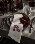 A styled dining table setting featuring a white linen napkin with an embroidered red monogram letter 'R' and floral border, layered over a dark metal charger plate. A red anthurium flower, a glass of red wine, a glass of water, and silver cutlery are arranged around it on a gray stone tabletop.