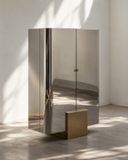 A tall, modern metallic cabinet with reflective silver doors and a matte gold base, photographed in a minimalist room with soft window shadows.