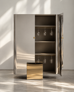 A modern mirrored double-door cabinet with a gold-toned interior and accessories, including hanging wine glasses and a matching gold side table, set against a minimalist wall with soft window shadows.