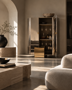 A tall mirrored cabinet with gold-toned hardware stands in a sunlit modern interior. The cabinet doors are open, showing shelves with wine bottles and hanging glassware, with a matching gold side table in front.
