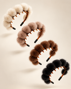 Four fluffy pom-pom headbands in cream, taupe, brown, and black, arranged diagonally on a warm beige background.