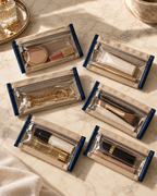 Six beige zippered cosmetic pouches with clear front panels and navy blue trim, arranged on a marble surface with makeup items inside each pouch.