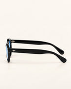 Black acetate sunglasses with a small silver logo on the temple arm, shown from a side profile view against a light cream background.