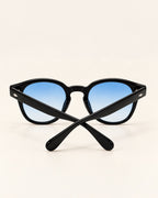 Black sunglasses with blue gradient lenses, shown crossed over each other on a light beige background.