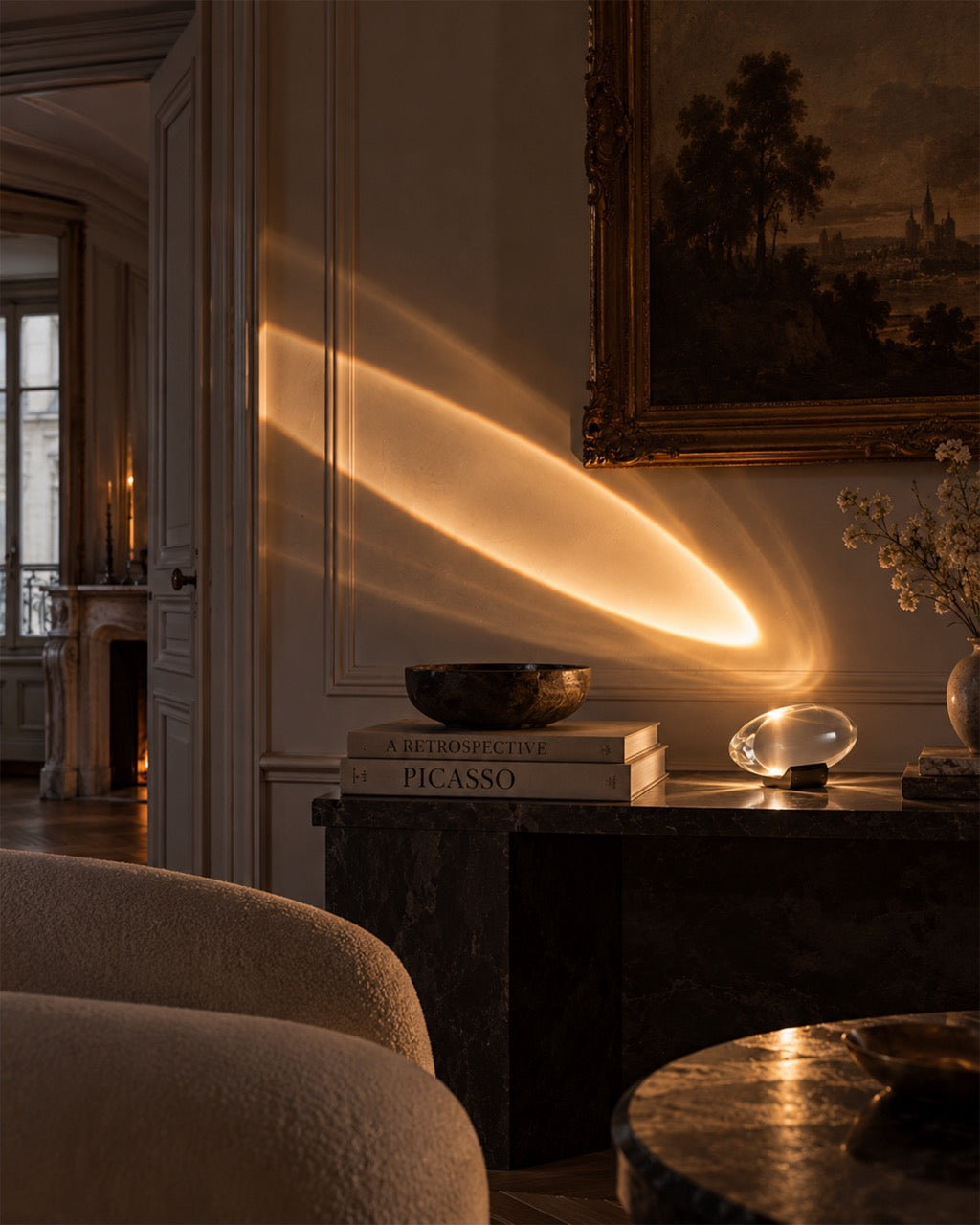 A warm, glowing oval light fixture casts a dramatic golden beam across a classical interior wall, with a framed painting, books, and furniture visible in the softly lit room.