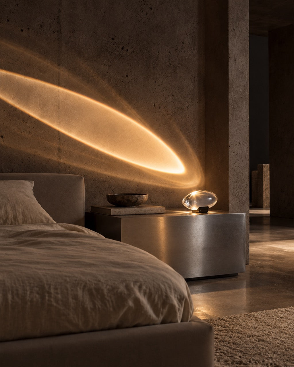 A modern bedroom scene with a low bed, concrete walls, and a sculptural floor lamp casting a warm oval beam of light across the wall. The lamp has a clear glass orb and a small black base, sitting on a metallic side table next to a bed with neutral bedding.