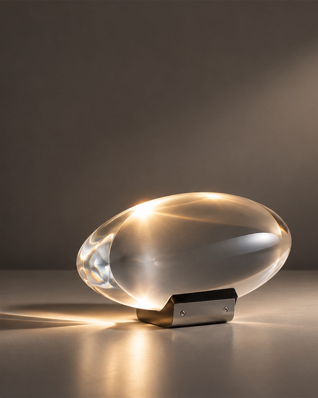 A modern oval-shaped translucent glass table lamp glowing with warm light, resting on a dark metal bracket against a dark gray background.