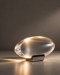 A modern oval-shaped translucent glass table lamp glowing with warm light, resting on a dark metal bracket against a dark gray background.