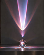 A clear glass crystal sphere projector sitting on a dark reflective surface, casting a dramatic cone of pink, purple, and blue light beams upward against a dark gray wall.