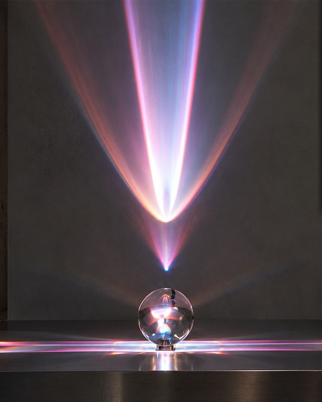 A clear glass crystal sphere projector sitting on a dark reflective surface, casting a dramatic cone of pink, purple, and blue light beams upward against a dark gray wall.